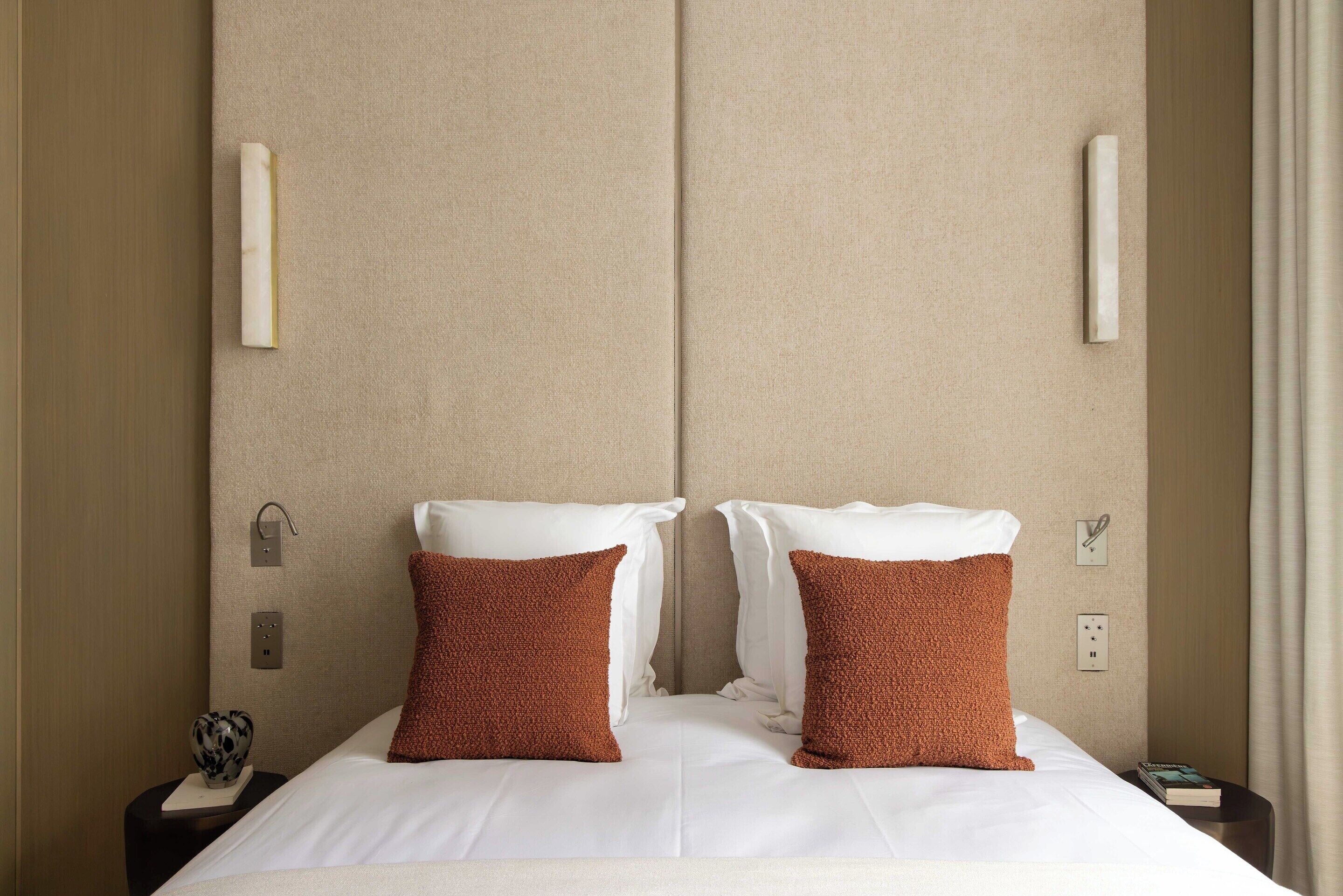Une chambre d’hôtel avec un grand lit, deux oreillers marron, un oreiller blanc et une table de chevet sur laquelle se trouvent un livre et un objet décoratif.