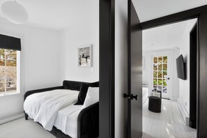 A modern bedroom with a bed, a framed picture, and a view of the outdoors. - Serene Designer Cottage Walking Distance to Town (Westhampton Beach)