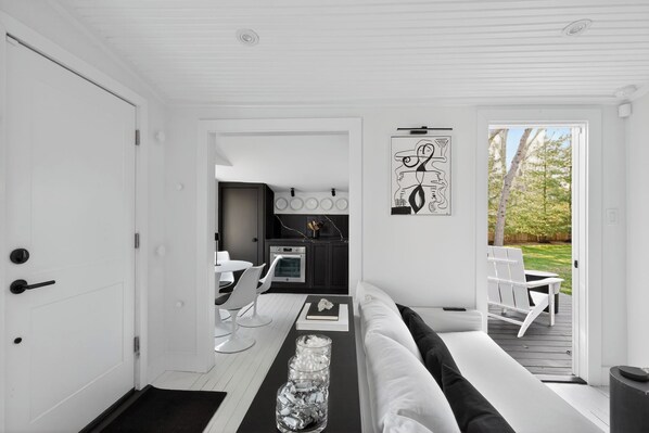 A modern interior with a white door, a kitchen area with a dining set, and a view through a glass door to an outdoor deck. - Serene Designer Cottage Walking Distance to Town (Westhampton Beach)