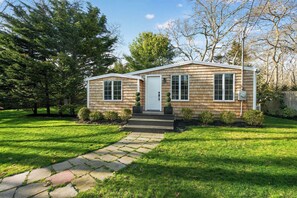 A single-story house with a front door, two windows on the front facade, a small porch with steps, and a stone pathway leading to the door. - Serene Designer Cottage Walking Distance to Town (Westhampton Beach)