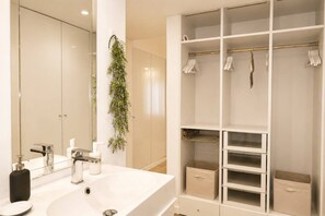 Ein modernes Badezimmer mit großem Spiegel, weißem Waschbecken und einem weißen begehbaren Kleiderschrank mit Regalen und Ablagefläche.