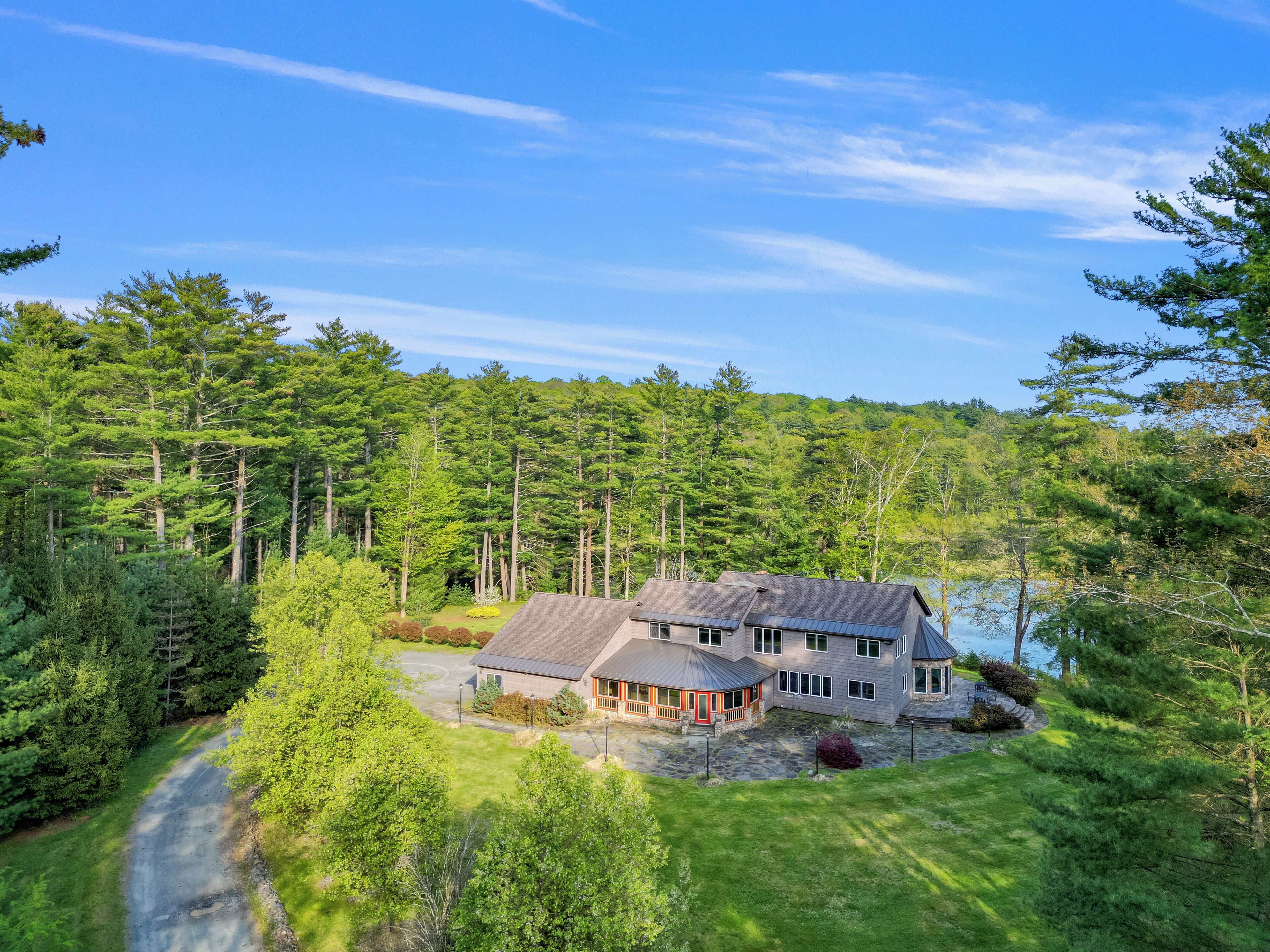 A large house with a stone driveway, surrounded by dense forest and a lake.
