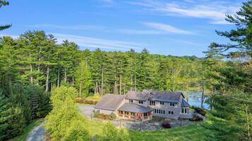 A large house with a stone driveway, surrounded by dense forest and a lake.