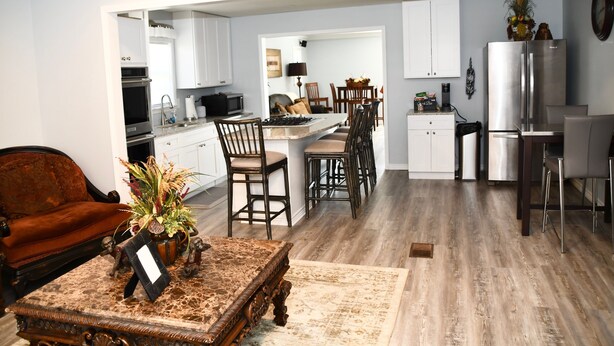 Una cocina moderna con armarios blancos, un comedor con sillas de madera y una sala de estar con sofá marrón y una mesa de centro.