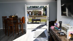 Una puerta abierta que da a una terraza con un banco, un comedor con sillas y una mesa, y un televisor montado en la pared.
