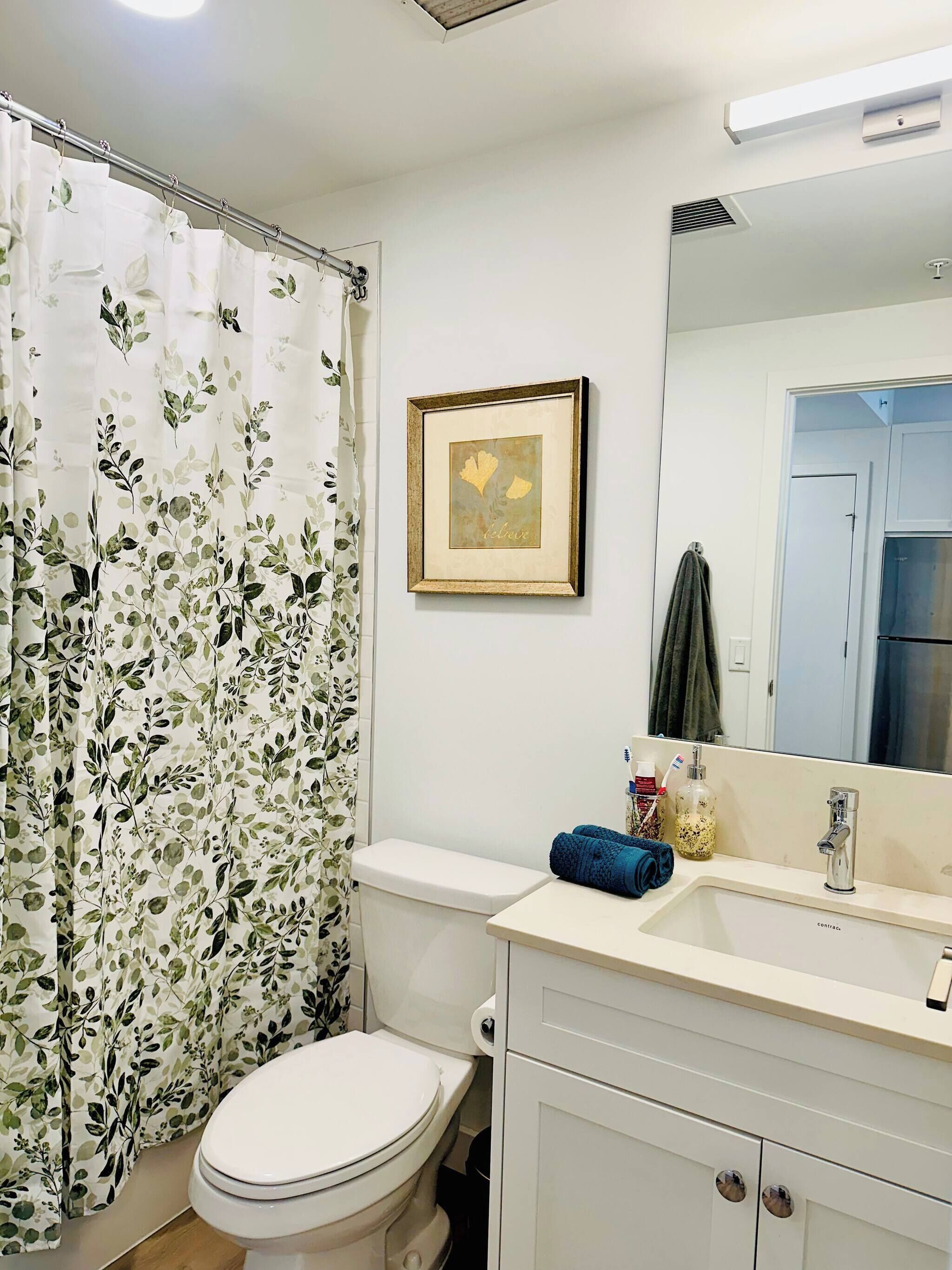 A bathroom with a toilet, a sink with a mirror, a framed picture on the wall, and a shower curtain with a leaf pattern.