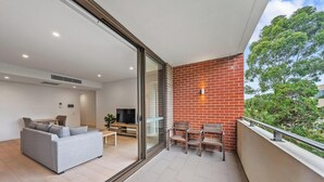 A balcony with a brick wall, a TV, a sofa, and a dining table. - KozyGuru | Roseville | Modern Leafy Retreat (Roseville)