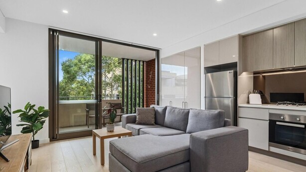 A modern living room with a grey sofa, a wooden coffee table, and a sliding glass door leading to a balcony. - KozyGuru | Roseville | Modern Leafy Retreat (Roseville)