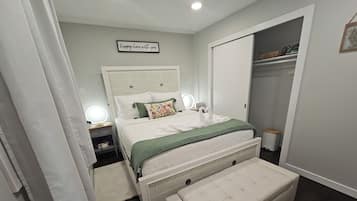 Un dormitorio con cama, lámparas de noche, una mesita de noche, un banco, un armario y un cartel en la pared.