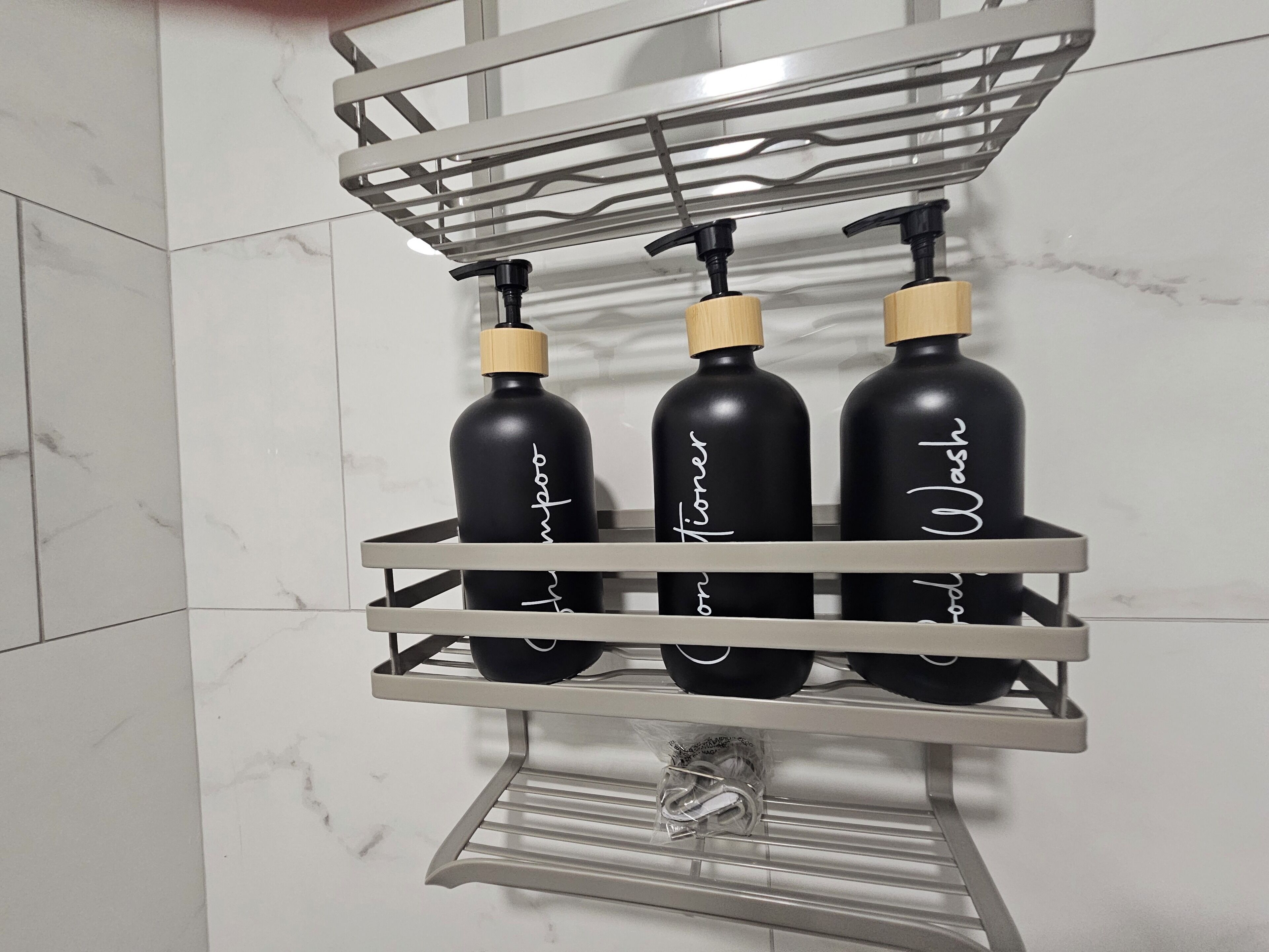 Estante de baño con tres botellas negras etiquetadas 'Ducha', 'Champú' y 'Acondicionador'.