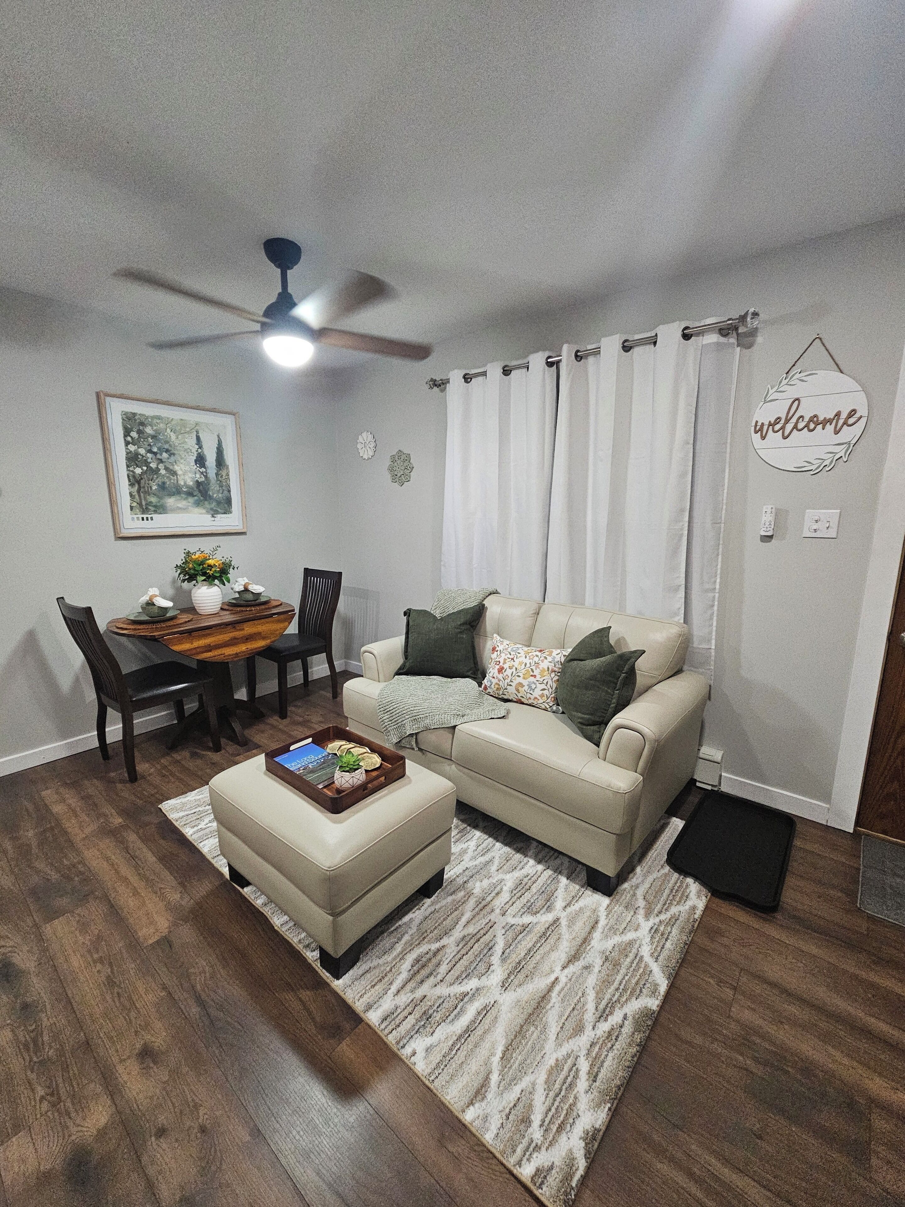 A living room with a sofa, a coffee table, a dining table with chairs, a ceiling fan, and a 'welcome' sign on the wall.