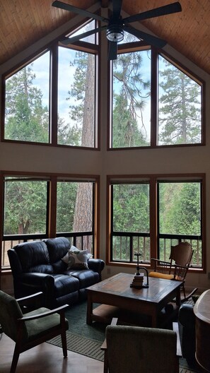 A spacious room with a high ceiling, large windows, a ceiling fan, a black leather sofa, a wooden coffee table, and a dining table with chairs. - Cedar Bend: Hilltop Secluded Retreat near Yosemite, Bass Lake & National Forest (Oakhurst)