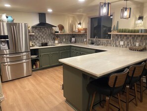 A modern kitchen with a stainless steel refrigerator, a large island with bar stools, and a tiled backsplash. -  Cedar Bend: Hilltop Secluded Retreat near Yosemite, Bass Lake & National Forest (Oakhurst)