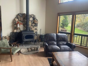 A stone fireplace, leather sofa, and a wooden coffee table in a living room with a view of a garden. -  Cedar Bend: Hilltop Secluded Retreat near Yosemite, Bass Lake & National Forest (Oakhurst)