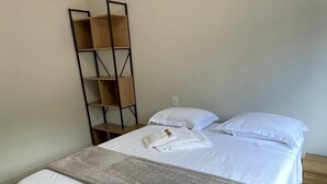 Una cama bien tendida con sábanas blancas y un plaid gris, una mesita de noche con cajón y una estantería metálica negra contra la pared.