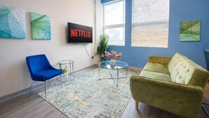 Ein moderner Büroraum mit blauer Wand, einem großen Bildschirm, der 'NETFLIX' anzeigt, einem blauen Sessel, einem gelben Sofa und abstrakten Kunstwerken.