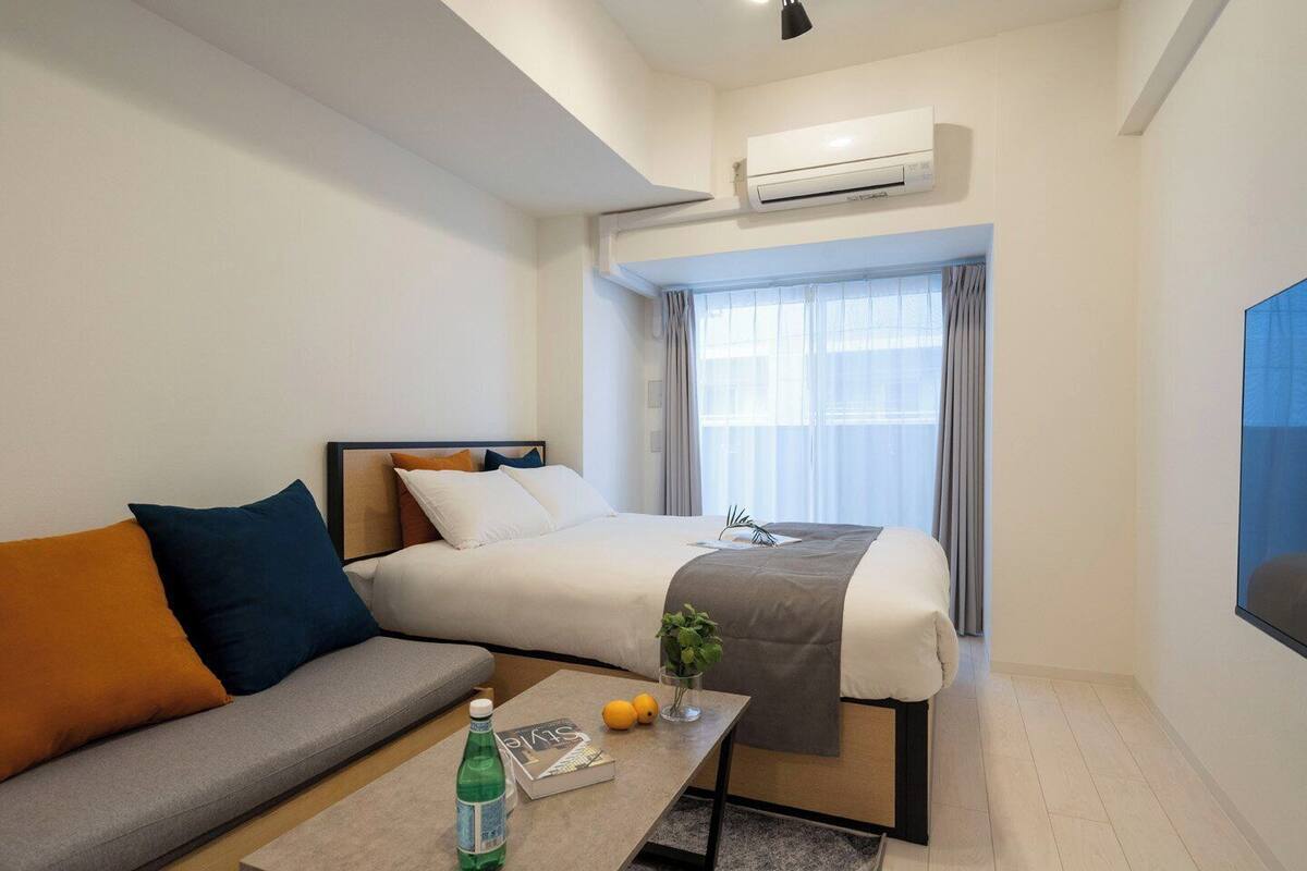 Una habitación de hotel con cama, sofá, una mesa de centro con una botella y un libro, aire acondicionado y ventana con cortinas.