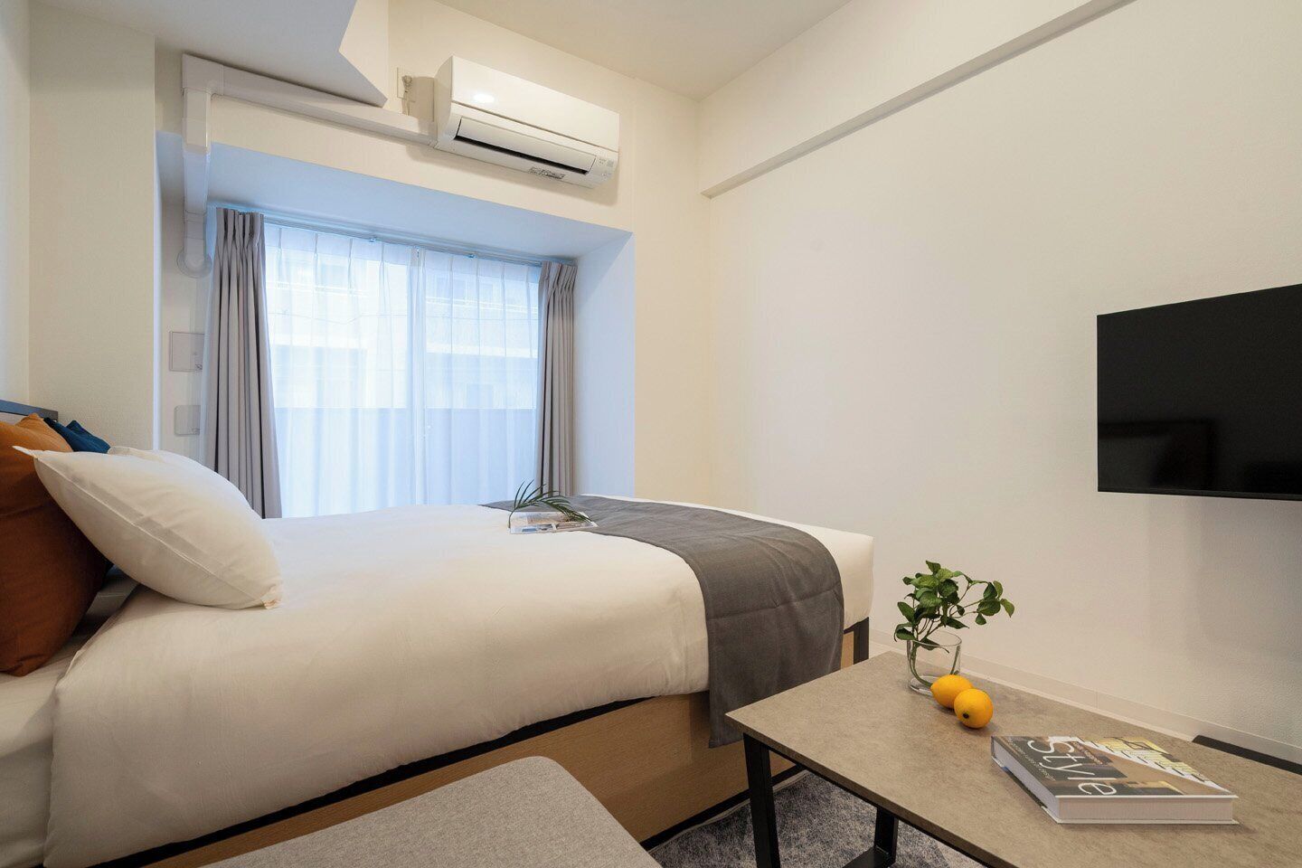 A hotel room with a bed, a flat-screen TV, a small table with a magazine, and a window with curtains.