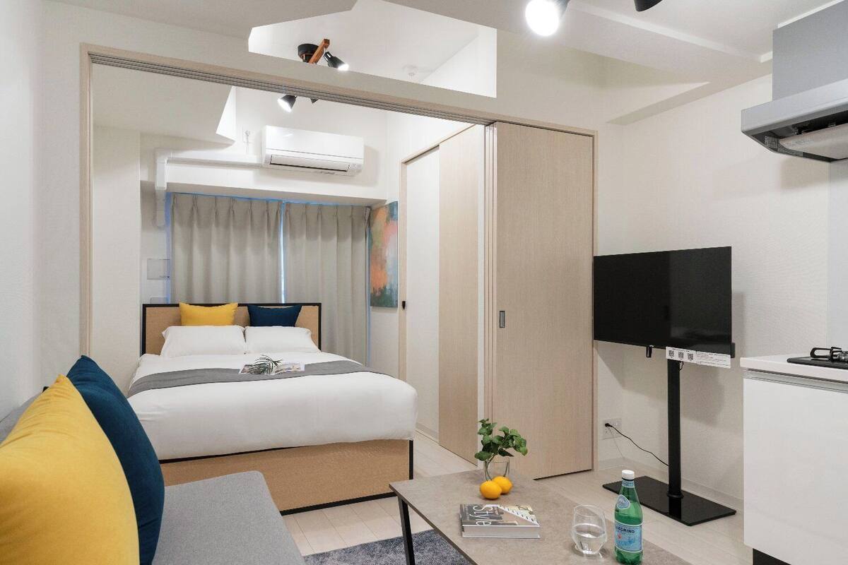 Una habitación de hotel moderna con una cama, un escritorio con televisor, una mesita con un libro y vasos, y un equipo de aire acondicionado.