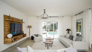 Un salon spacieux avec une grande télévision, une étagère en bois, un canapé, une table basse et une vue sur la piscine, visible à travers les portes vitrées.