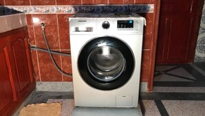 Samsungin pesukone keittiössä, jossa on laatoitetut seinät ja puuovi.