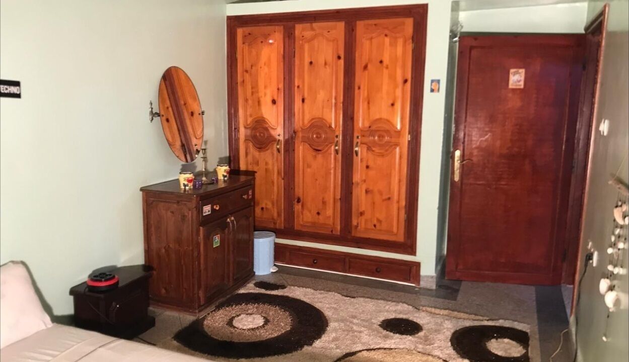 A room with a wooden wardrobe, a small table with a mirror, a bed, and a door.