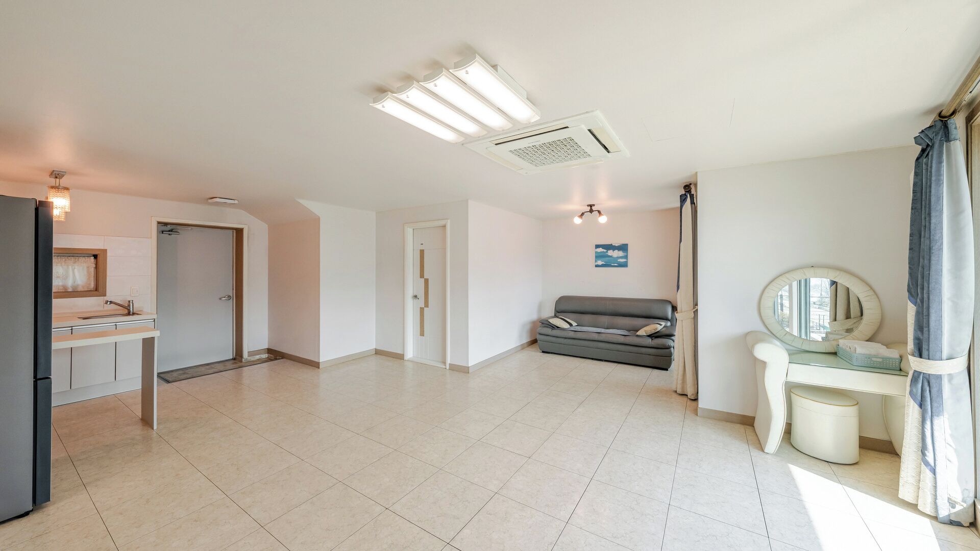 A spacious living area with a kitchenette, a gray sofa, a small table with a mirror, and a ceiling-mounted air conditioning unit.