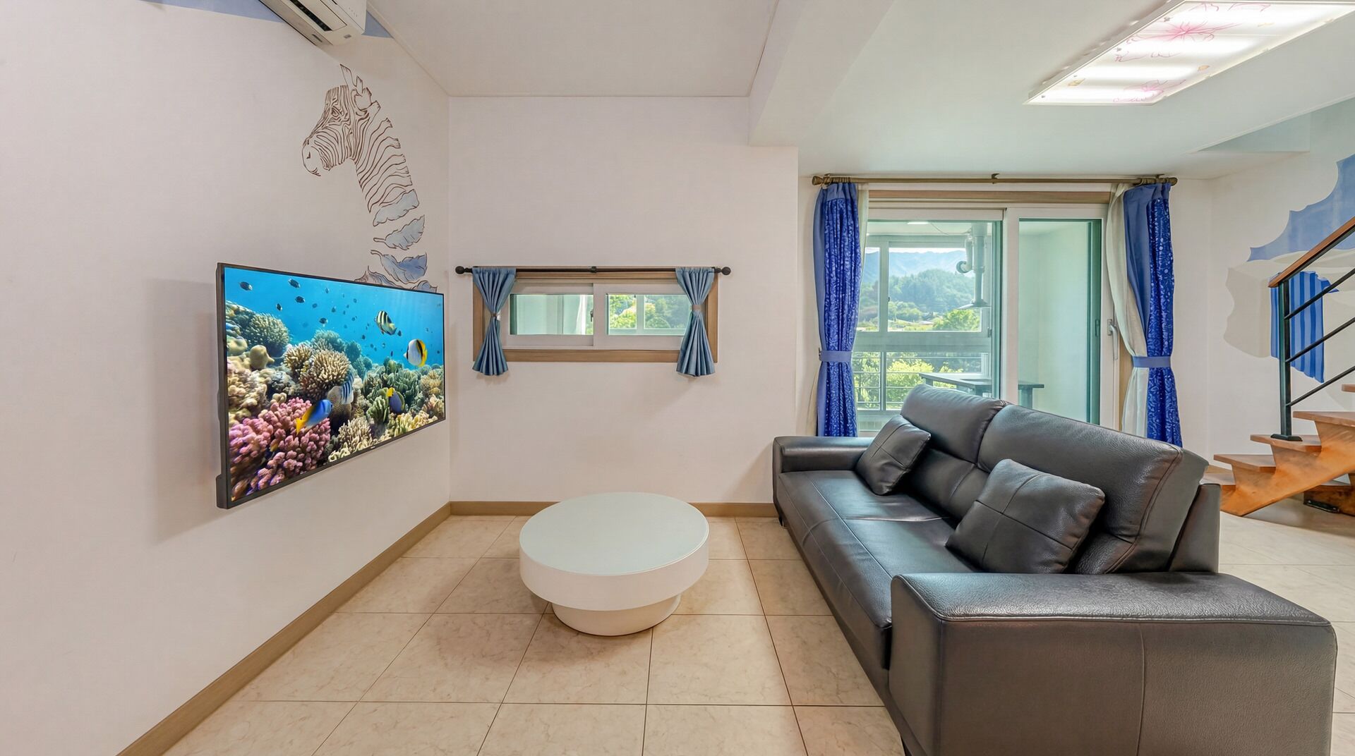 A living room with a sofa, a round coffee table, a wall-mounted TV, and a window with curtains.