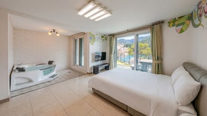 A hotel room with a bed, a TV, a bathtub, and a balcony with a view of mountains and cherry blossoms.