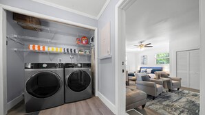 Un cuarto de lavandería con dos lavadoras de carga frontal, repisas con productos de limpieza y vista a una sala de estar con sofá y ventilador de techo.