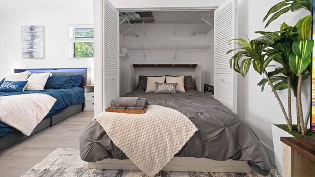 Un dormitorio con cama, mesita de noche, una silla, una planta y una ventana.