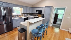 A kitchen with grey cabinets, a stainless steel refrigerator, a microwave, and a sink. There are three bar stools and a small island with a white countertop. - Stone Landing Lakefront Retreat: Spacious 2-bed, 3-bath House with Private Dock (Fair Play)