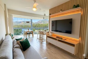 A modern living room with a large TV, a sofa, a dining area, and a view of mountains. - Encanto da Praia Luxo  (Niterói)