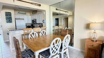 A dining area with a wooden table and white chairs, a kitchen in the background, and a mirror reflecting the dining area.