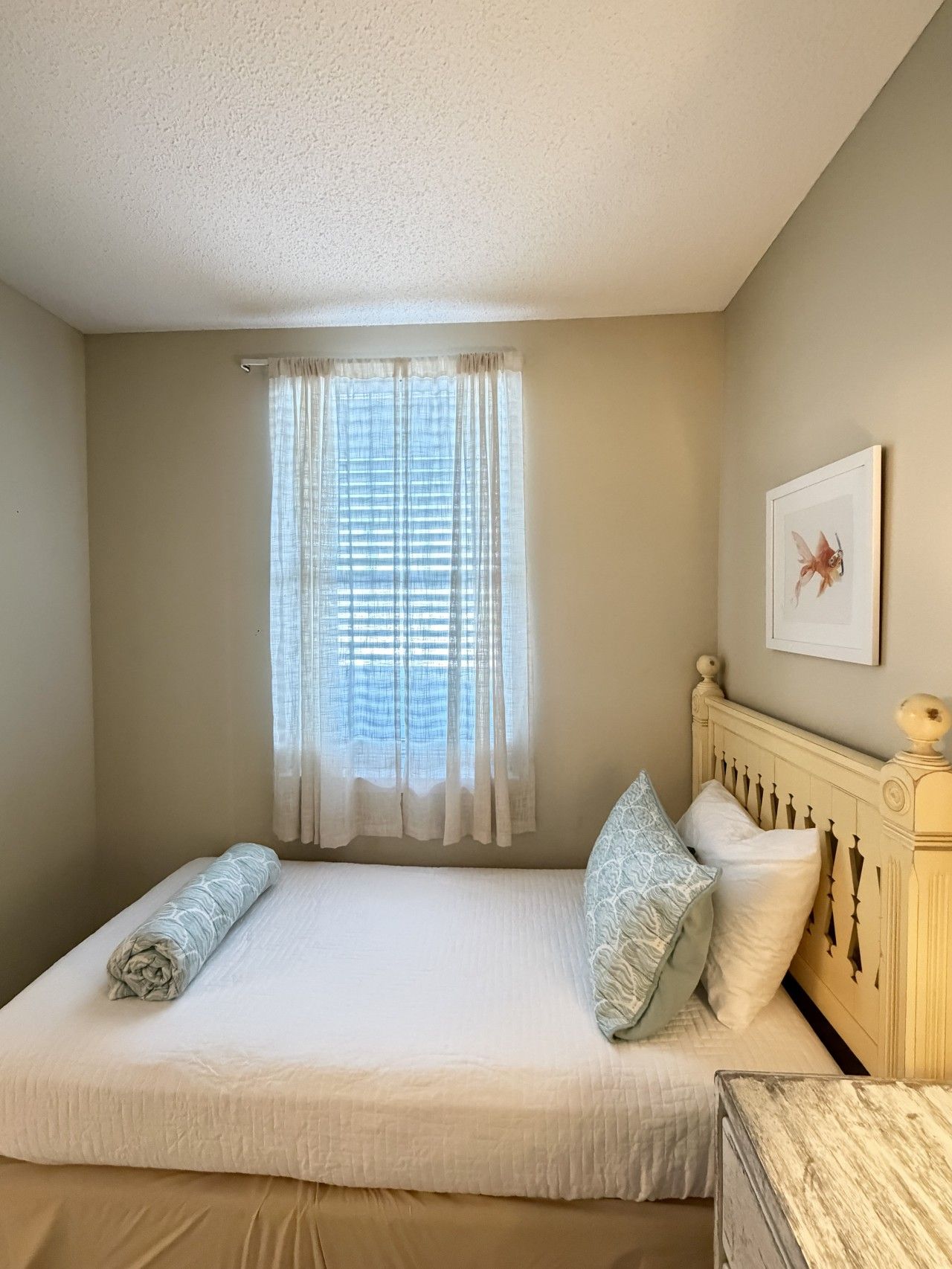 A bedroom with a bed, two pillows, a window with curtains, and a framed picture on the wall.