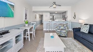 A modern living room with a flat-screen TV, a ceiling fan, a dining area with chairs, and a kitchen in the background. - Sundestin 105 (Destin)