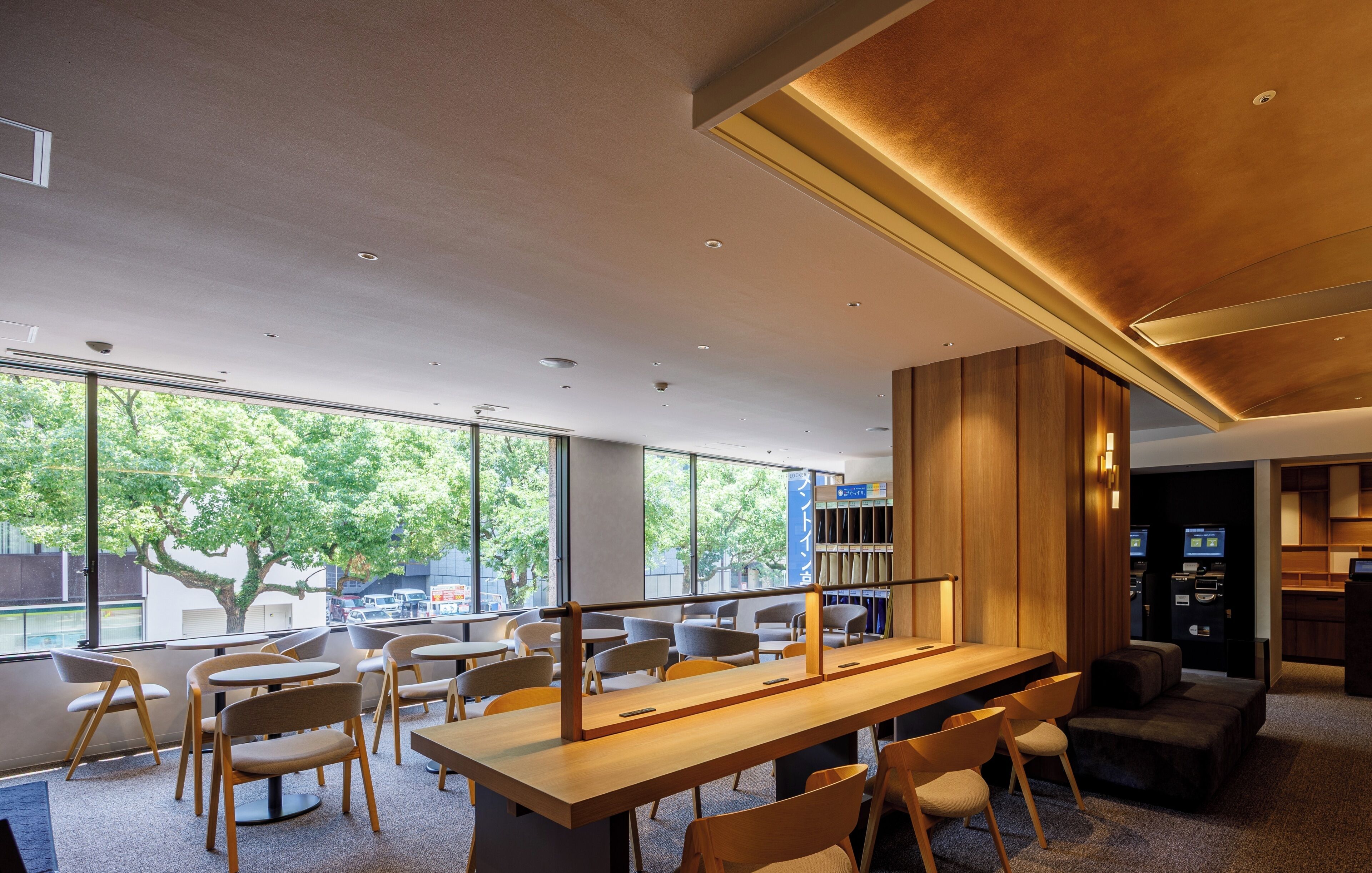 Un intérieur moderne avec une longue table en bois, de nombreuses chaises et de grandes fenêtres donnant sur les arbres et les bâtiments environnants.