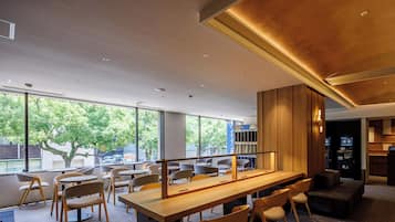 A modern interior with a long wooden table, multiple chairs, and large windows offering a view of trees and buildings outside.