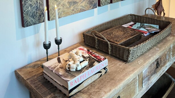 Een houten console tafel met een boek getiteld 'RALPH LAUREN', een kandelaar en een mand met tijdschriften.