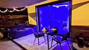 Una habitación con puerta de vidrio, una mesa pequeña con sillas, un sofá y una butaca.