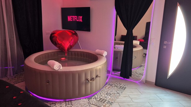 Una habitación con una bañera de hidromasaje circular, un globo con forma de corazón, un cartel de Netflix y un espejo que refleja la habitación.