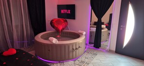 Ein Zimmer mit einer runden Whirlwanne, einem herzförmigen Luftballon, einem Netflix-Schild und einem Spiegel, der das Zimmer reflektiert.