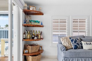 A room with a grey sofa, a wooden shelf unit with bottles and baskets, and a window with shutters. - Waterfront 2BR w/ Canal, Patio & Optional Dockage – Tavernier Keys Getaway (Marathon)