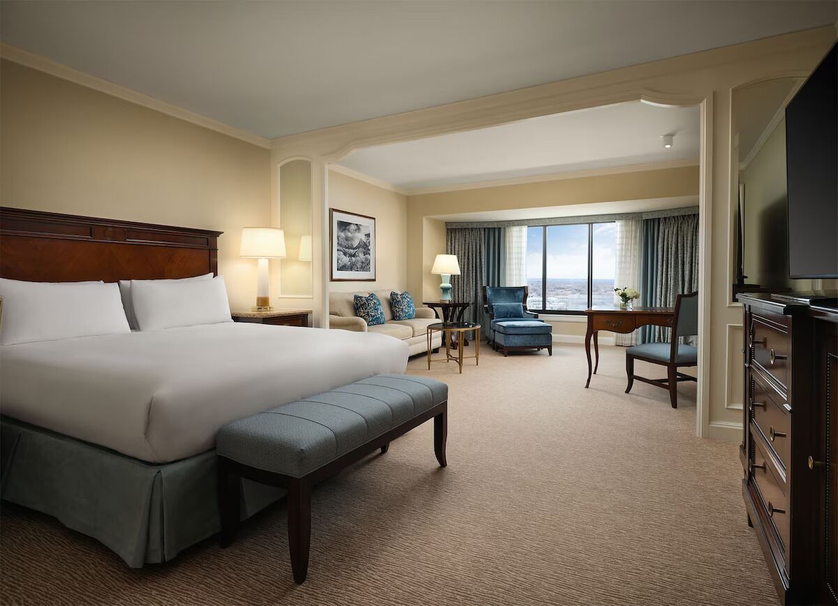 A hotel room with a large bed, a desk, a chair, a TV, and a view of the city.