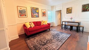 En stue med en rød sofa, et mønstret gulvtæppe, et spisebord med bænke og indrammede kunstværker på væggene.