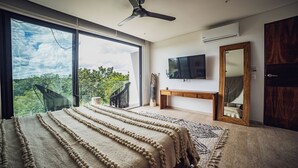 A bedroom with a large bed, a ceiling fan, a TV, and a view of greenery. - Selvatico PH and private pool by BT Homes (Tulum)