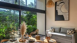 Un coin repas avec une table dressée pour quatre personnes, un canapé agrémenté de coussins à motifs et une grande fenêtre donnant sur un jardin luxuriant.