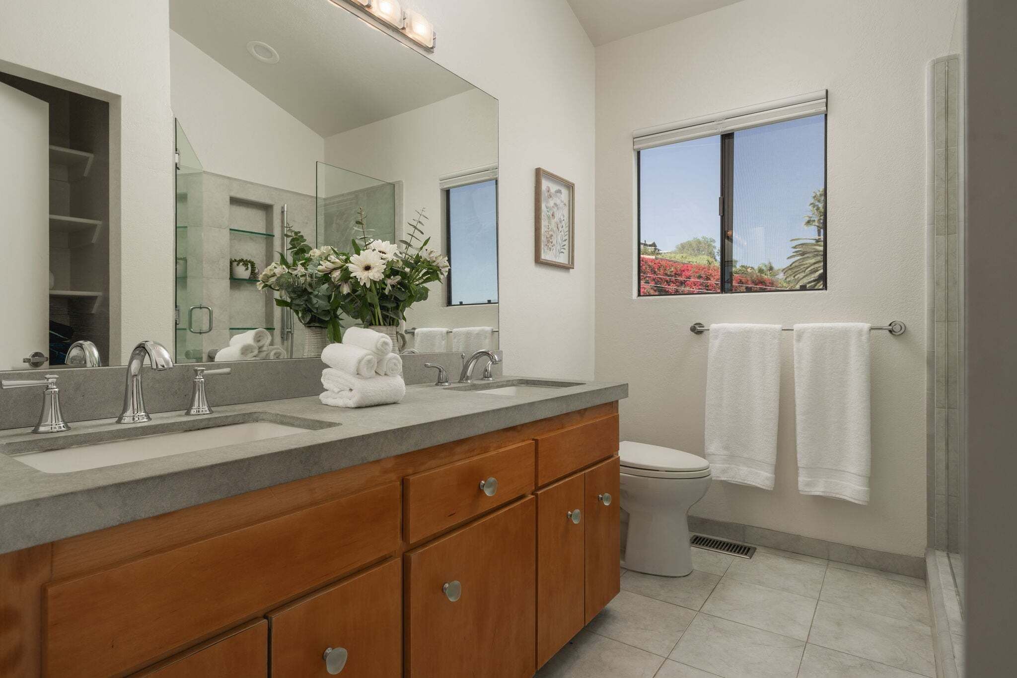 A modern bathroom with a double sink, wooden vanity, large mirror, and a window with a view of red flowers.