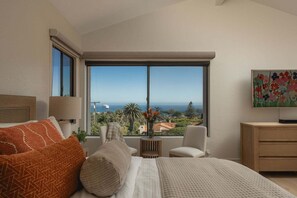 Ein Schlafzimmer mit großem Fenster, einem Bett, einem Sessel, einem kleinen Tisch und einem Gemälde.