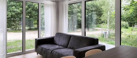 A modern living room with a dark grey sofa, a wooden dining table, and large windows offering a view of a green landscape.
